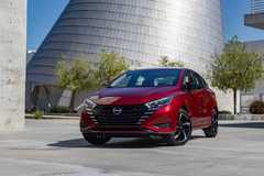 Front 3/4 view of a red 2025 Nissan Versa with a modern glass building in the background.