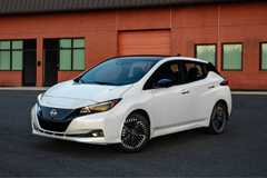 Front 3/4 view of a white 2025 Nissan Leaf parked on tarmac outside of a brick building.