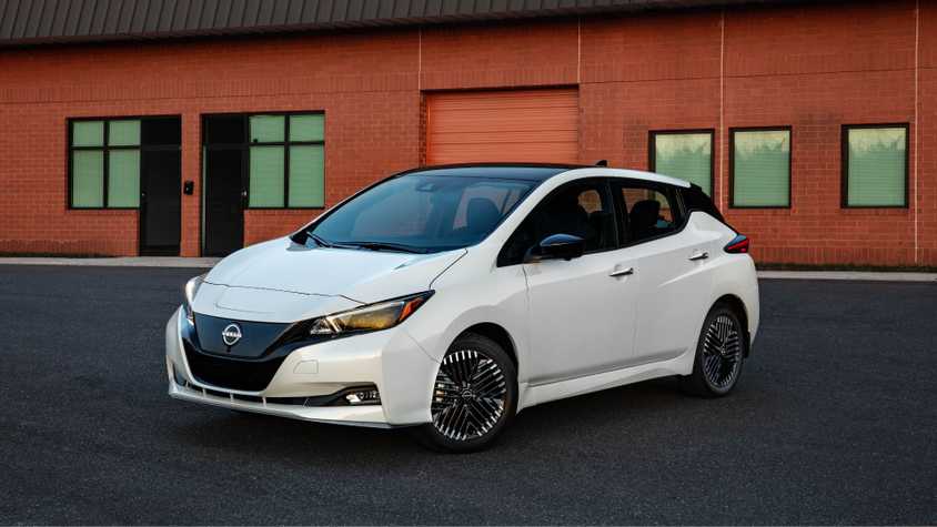 Front 3/4 view of a white 2025 Nissan Leaf parked on tarmac outside of a brick building.