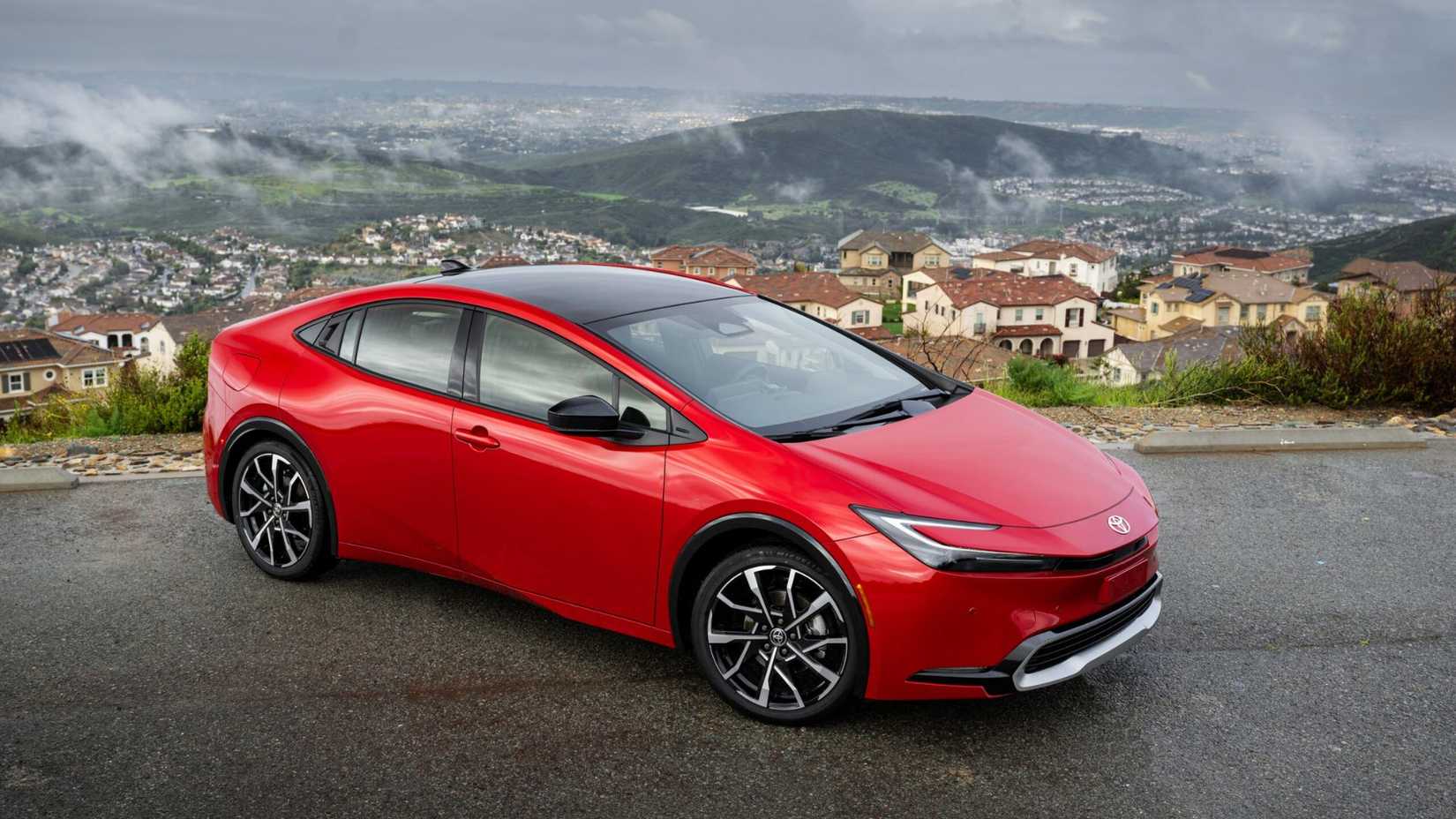 Static front 3/4 shot of a red 2025 Toyota Prius PHEV XSE with clouds and a town in the background.