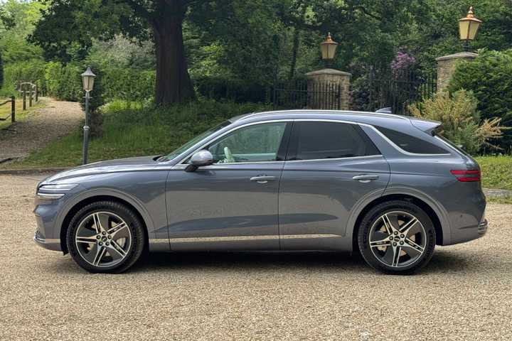 Static side profile shot of a gray 2026 Genesis Electrified GV70 parked on gravel with trees in the background.