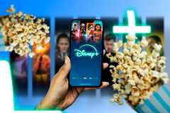 A hand holding a phone with the Disney+ screen displayed, and two popcorn bags next to it.