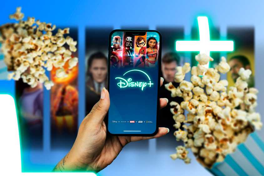 A hand holding a phone with the Disney+ screen displayed, and two popcorn bags next to it.