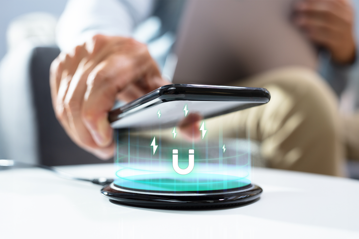 A hand lowers a smartphone onto a wireless charging pad, with glowing magnetic energy lines and a magnet icon projected between them.