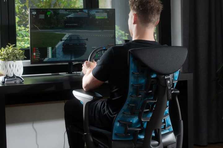 A man plays a racing game at a desk using a Logitech G29 racing wheel.