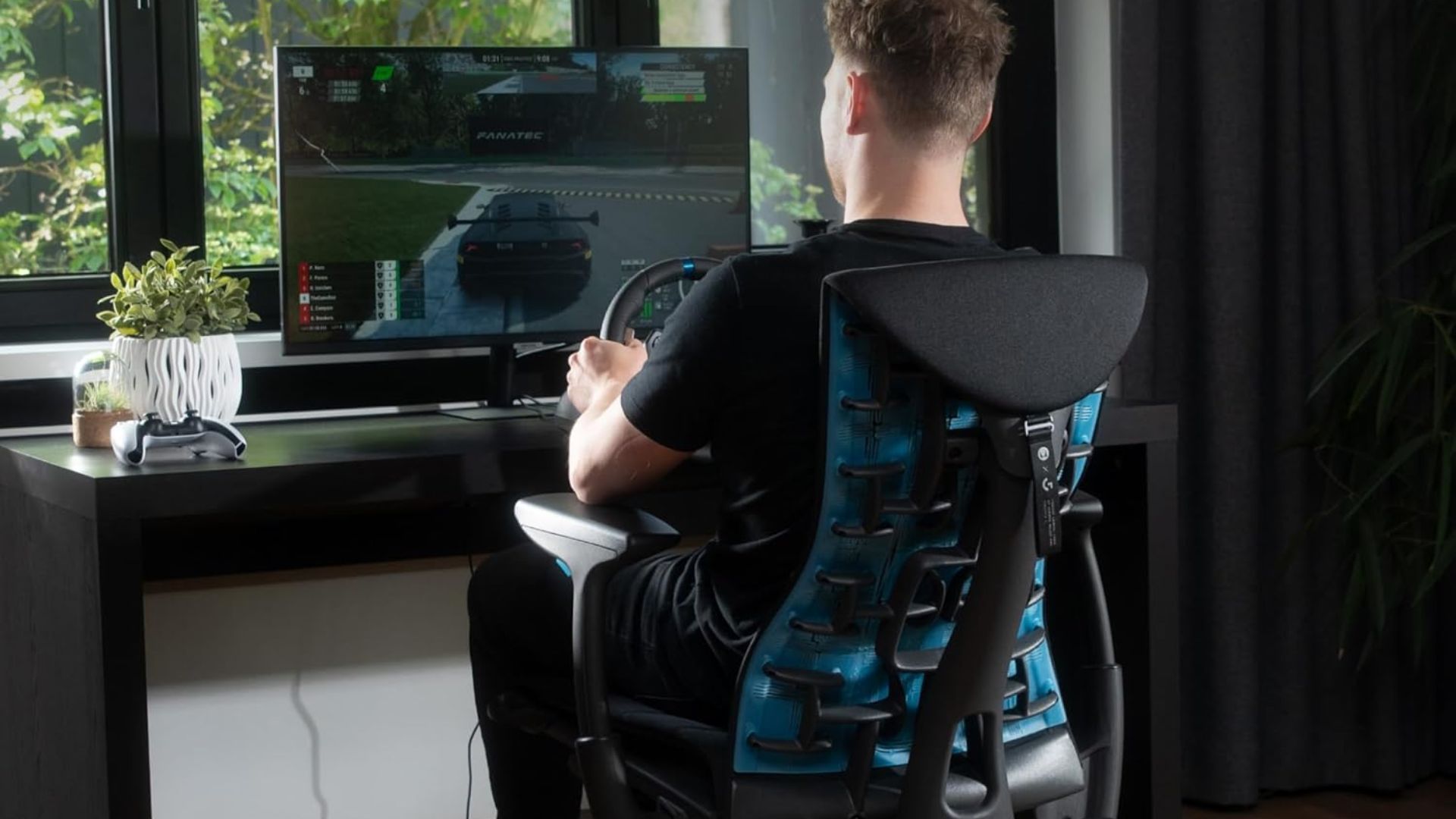 A man plays a racing game at a desk using a Logitech G29 racing wheel.