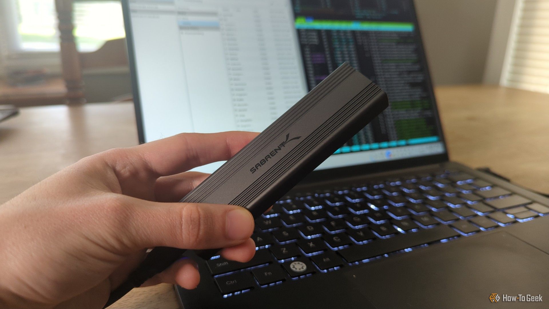 A person's hand holding a Sabrent external SSD drive in front of a Linux laptop.