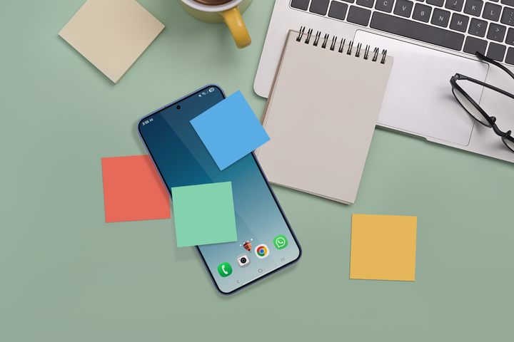 A smartphone with colorful sticky notes placed on its screen lies on a desk, surrounded by a notepad, glasses, a laptop, and a coffee cup.