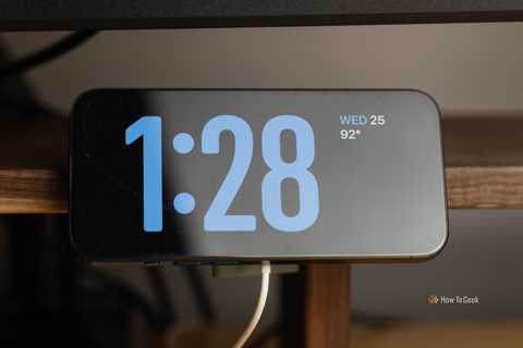 An iPhone mounted in Standby Mode on a 3D printed MagSafe charger mount that is clamped to a walnut desk shelf.