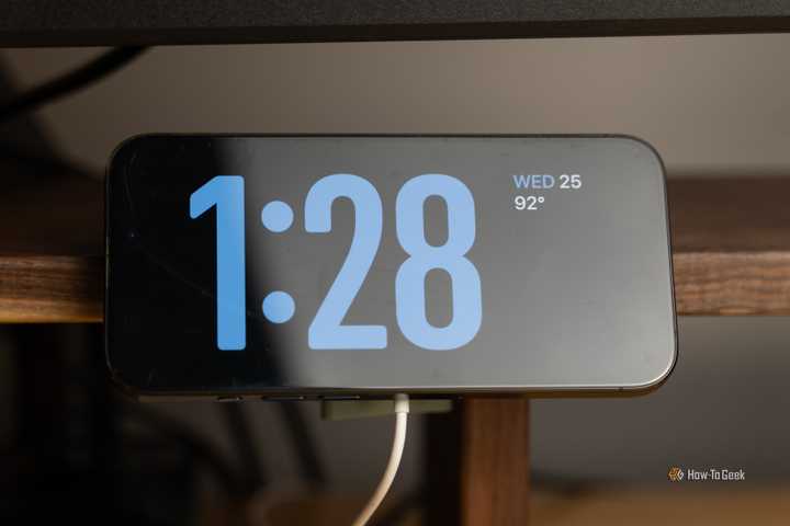 An iPhone mounted in Standby Mode on a 3D printed MagSafe charger mount that is clamped to a walnut desk shelf.
