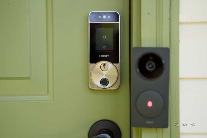 The Lockly Visage smart lock mounted on a green front door with a black doorbell to its right.