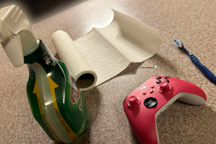 A Pink Xbox One Controller with a bottle of cleaner, a toothbrush, a roll of paper towels, and a metal pick.