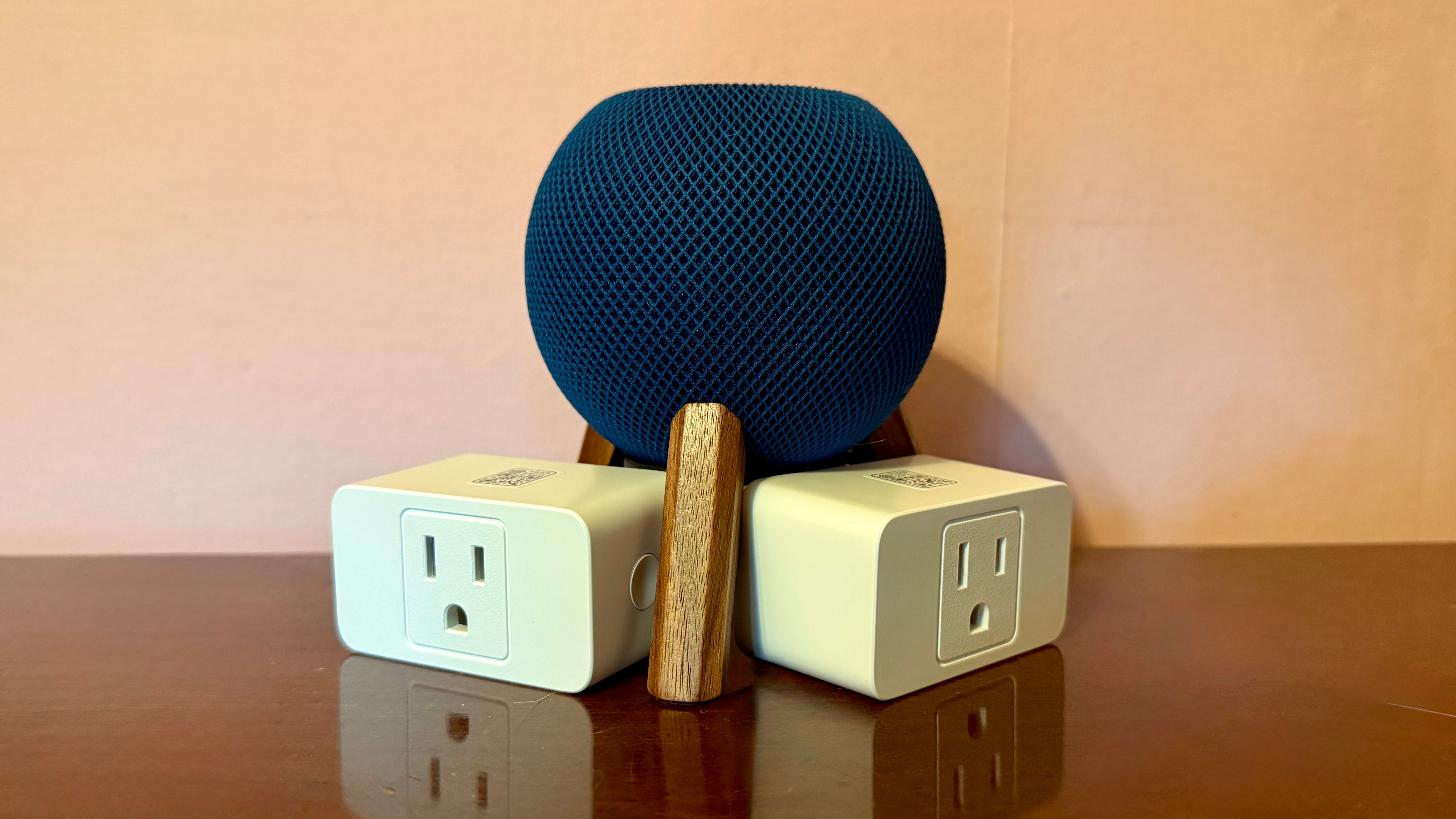 HomePod mini surrounded by smart plugs and on a wooden stand on top of a wooden surface.