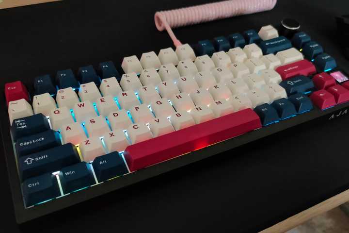 The Ajazz AK820 Pro mechanical keyboard with a pink coiled cable and white, blue, and red keycaps sitting on a desk with a black SteelSeries mousepad.