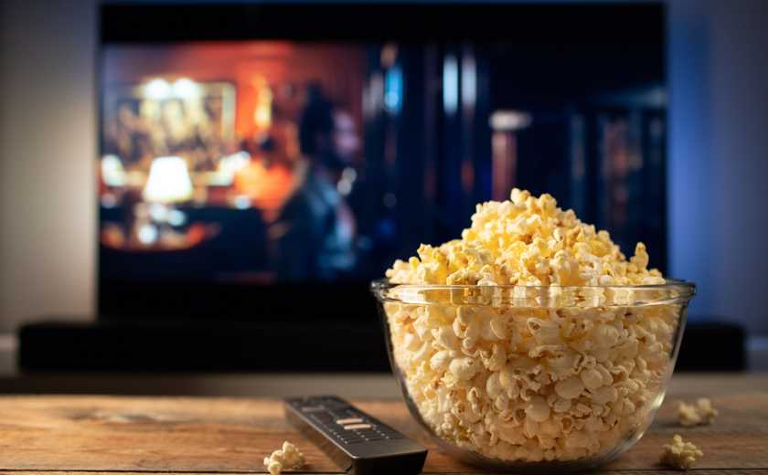 A glass bowl of popcorn and remote control while in the background the TV plays.
