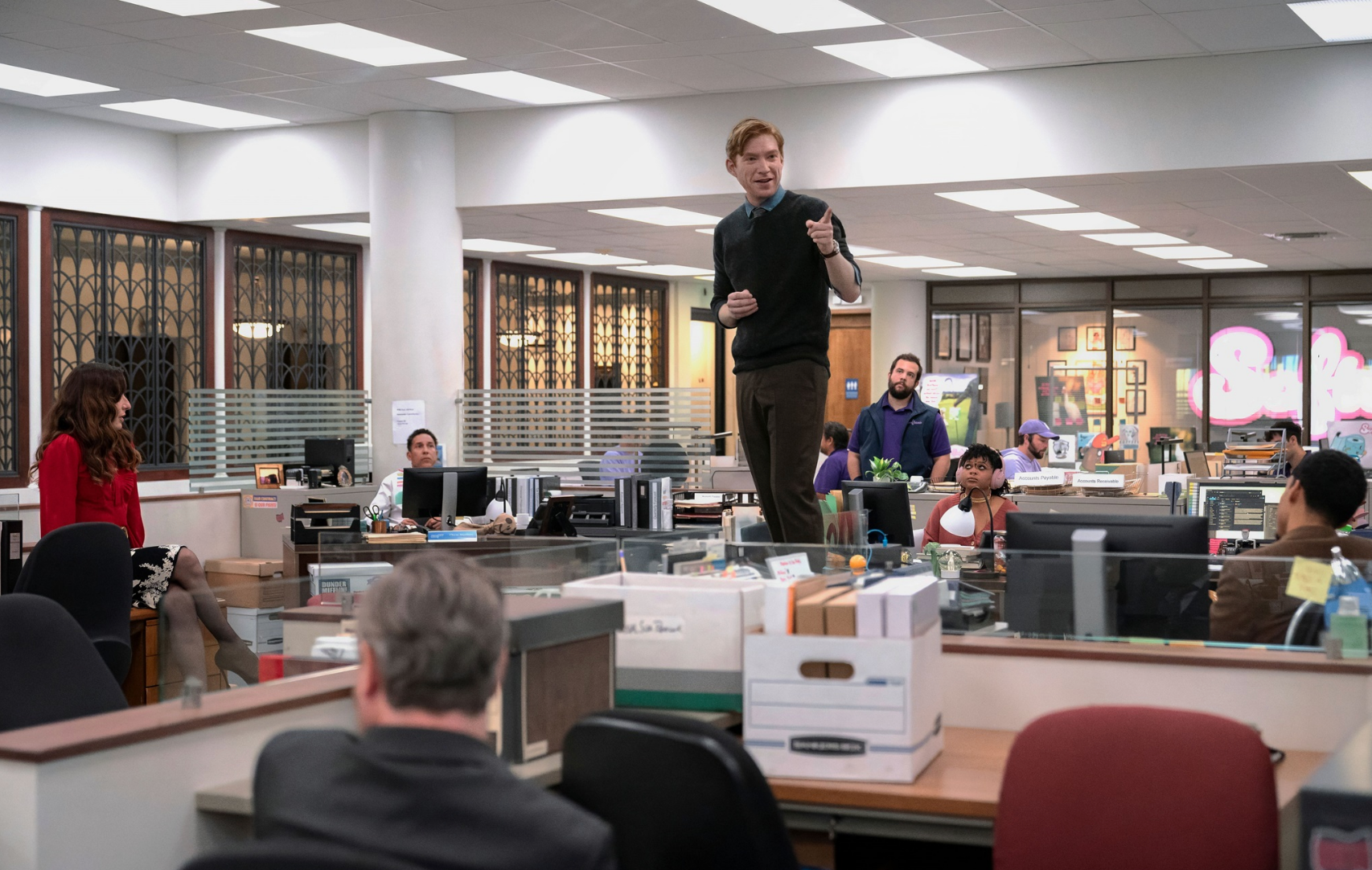 Domhnall Gleeson standing atop a desk talking to his staff.