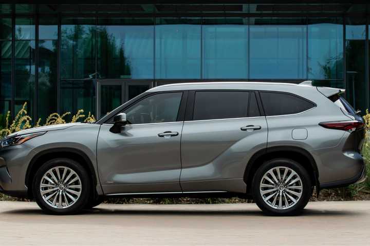 Static side profile shot of a gray 2025 Toyota Highlander Hybrid parked outside of a modern glass-fronted building.