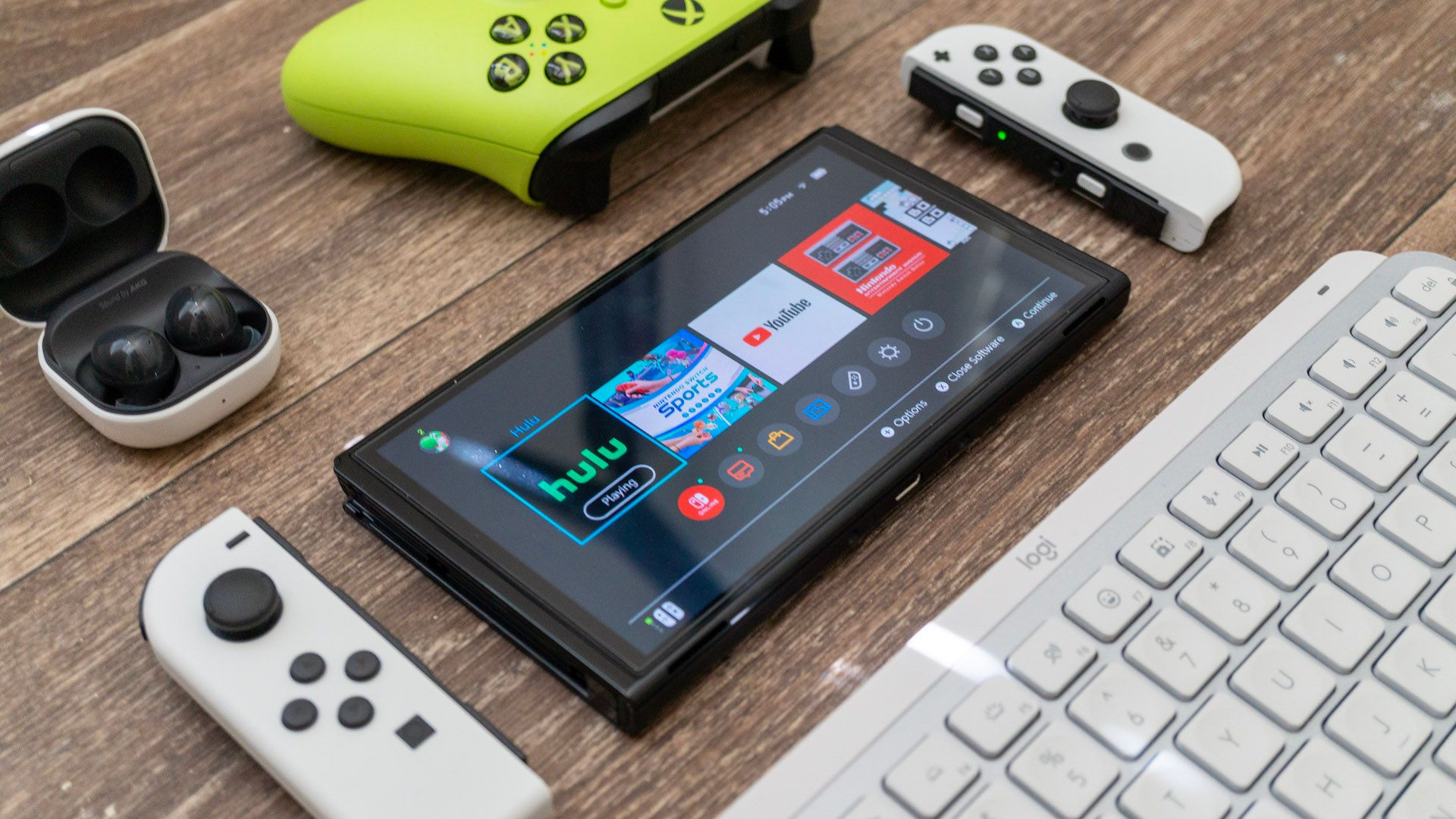 Nintendo Switch laying on a table next to a keyboard, Samsung earbuds, and an Xbox controller