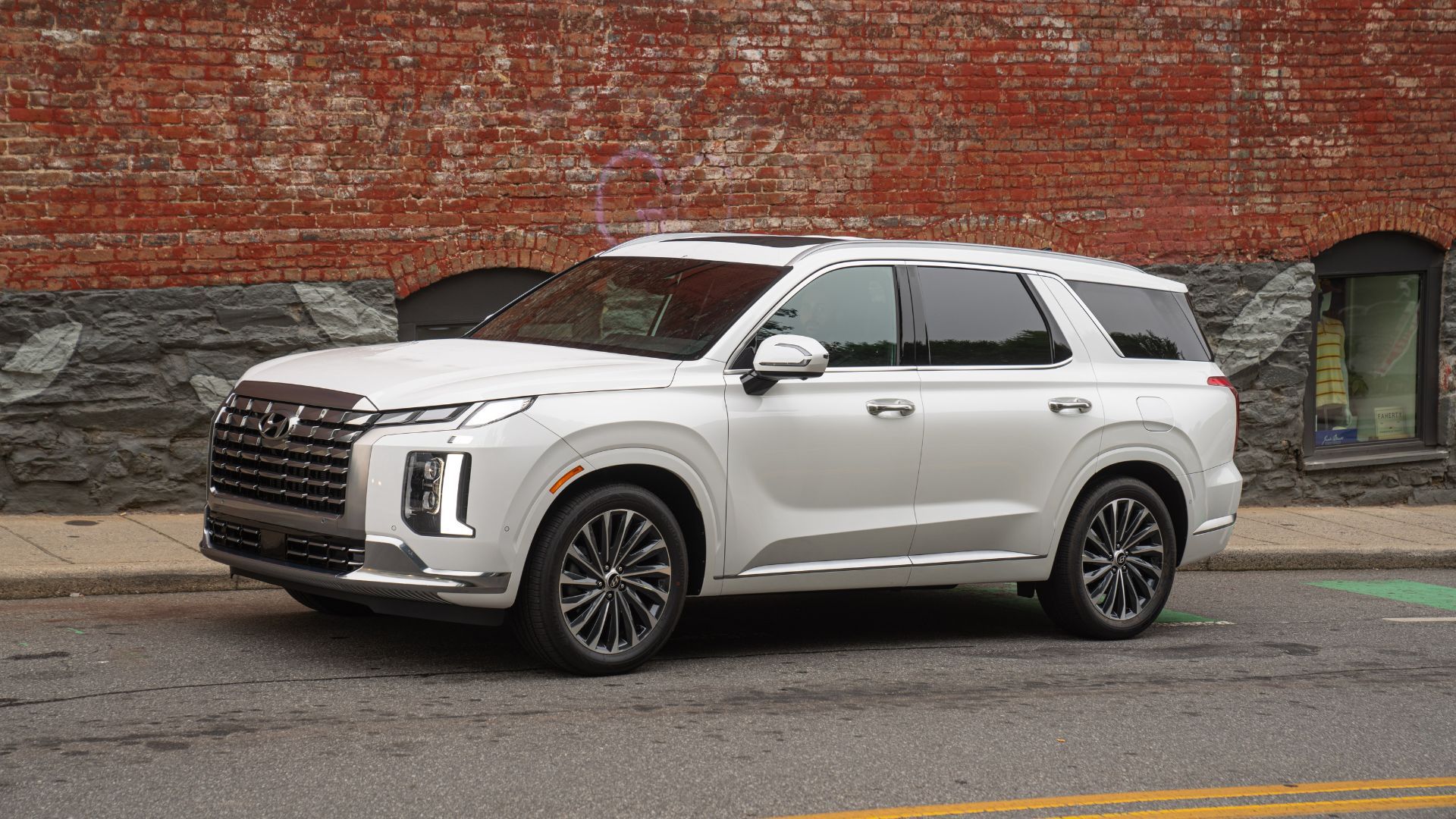 Front three-quarter view of a white 2025 Hyundai Palisade parked alongside a sidewalk with a brick wall with graffiti on it.