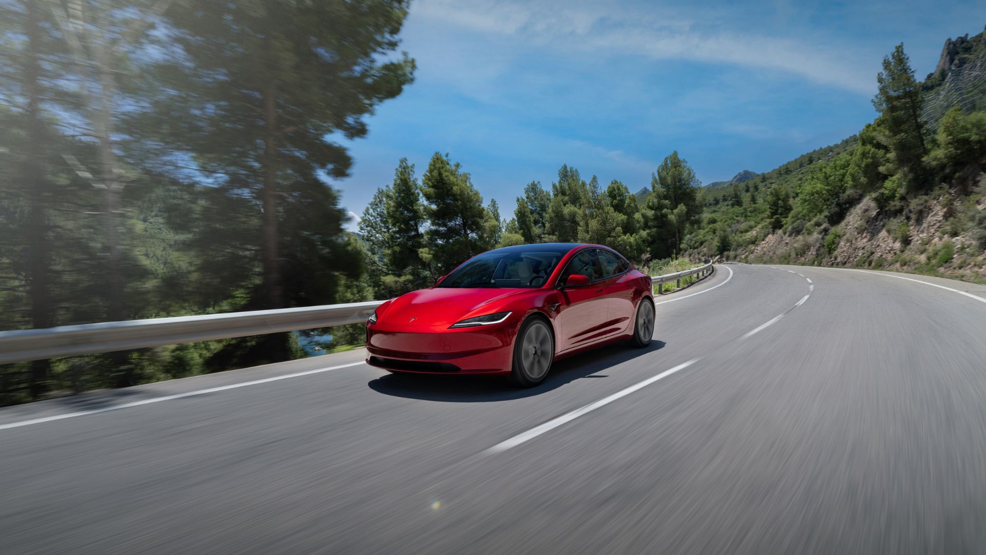 Front 3/4 shot of a red 2024 Tesla Model 3 driving on a winding country road.