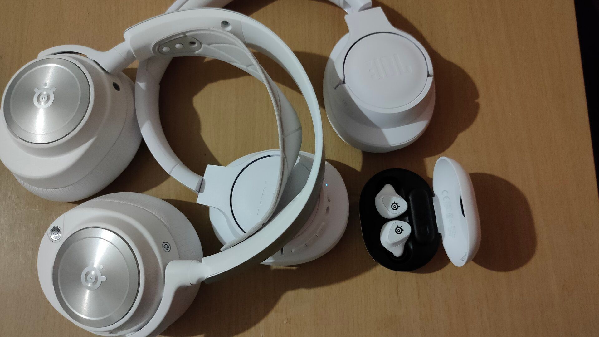 White SteelSeries Arctis Nova Pro Wireless headset, SteelSeries Arctis GameBuds, and JBL 770NC active noise-cancelling headphones placed on a desk.