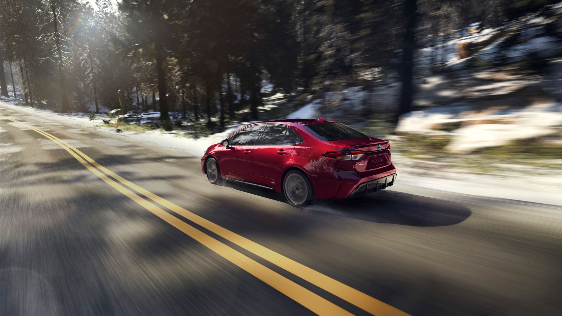Dynamic rear 3/4 shot of a red 2025 Toyota Corolla Hybrid driving on a country road with trees lining the road.