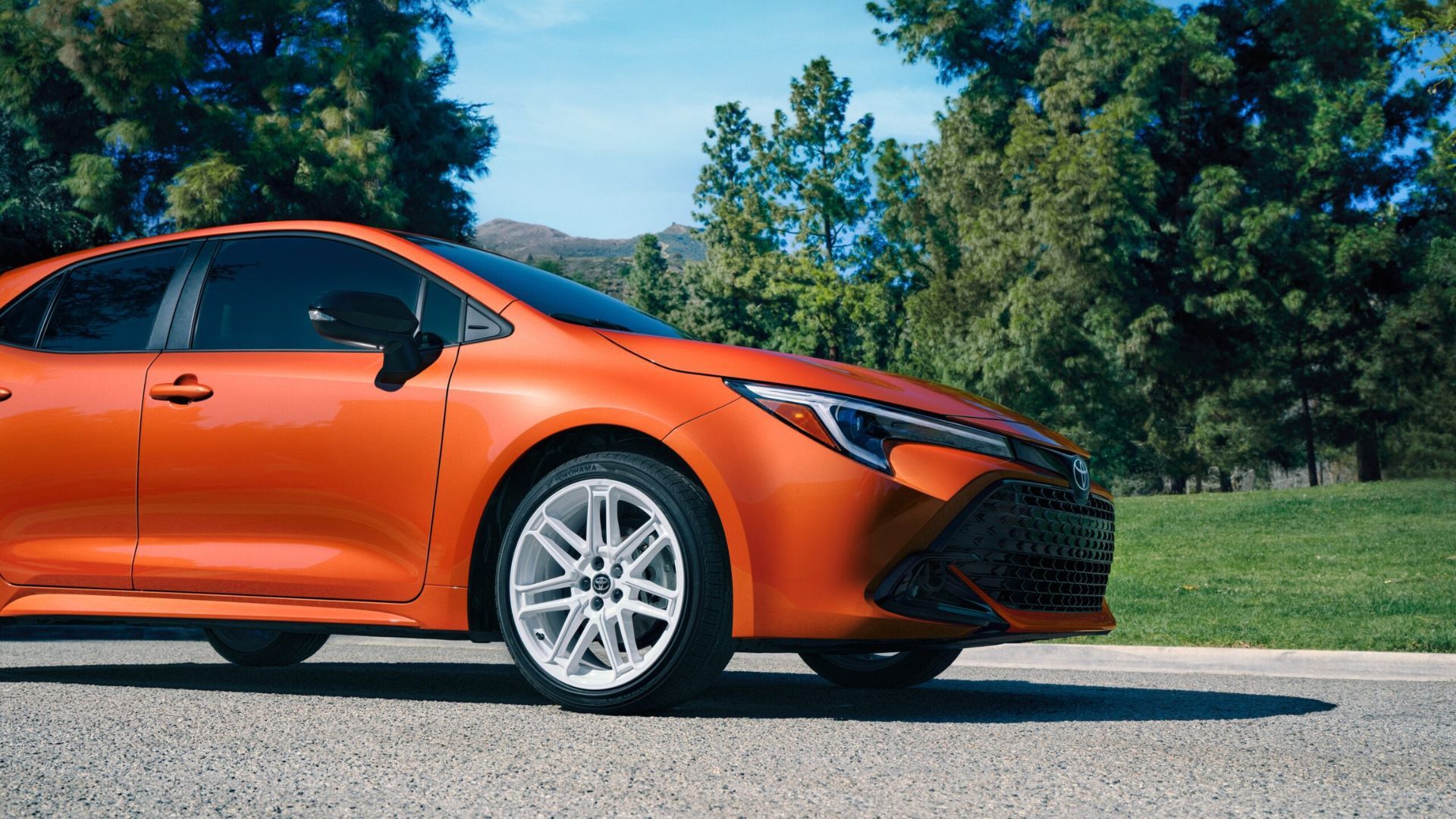 Static shot of the front end of an orange 2026 Corolla Hatchback FX Edition with trees in the background.