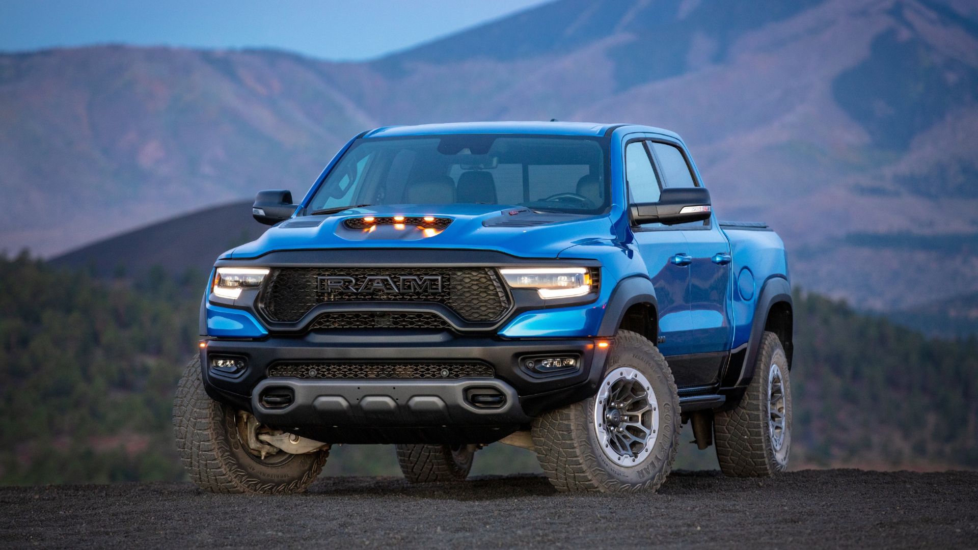 Static front 3/4 shot of a blue 2024 Ram 1500 TRX with mountains in the background.