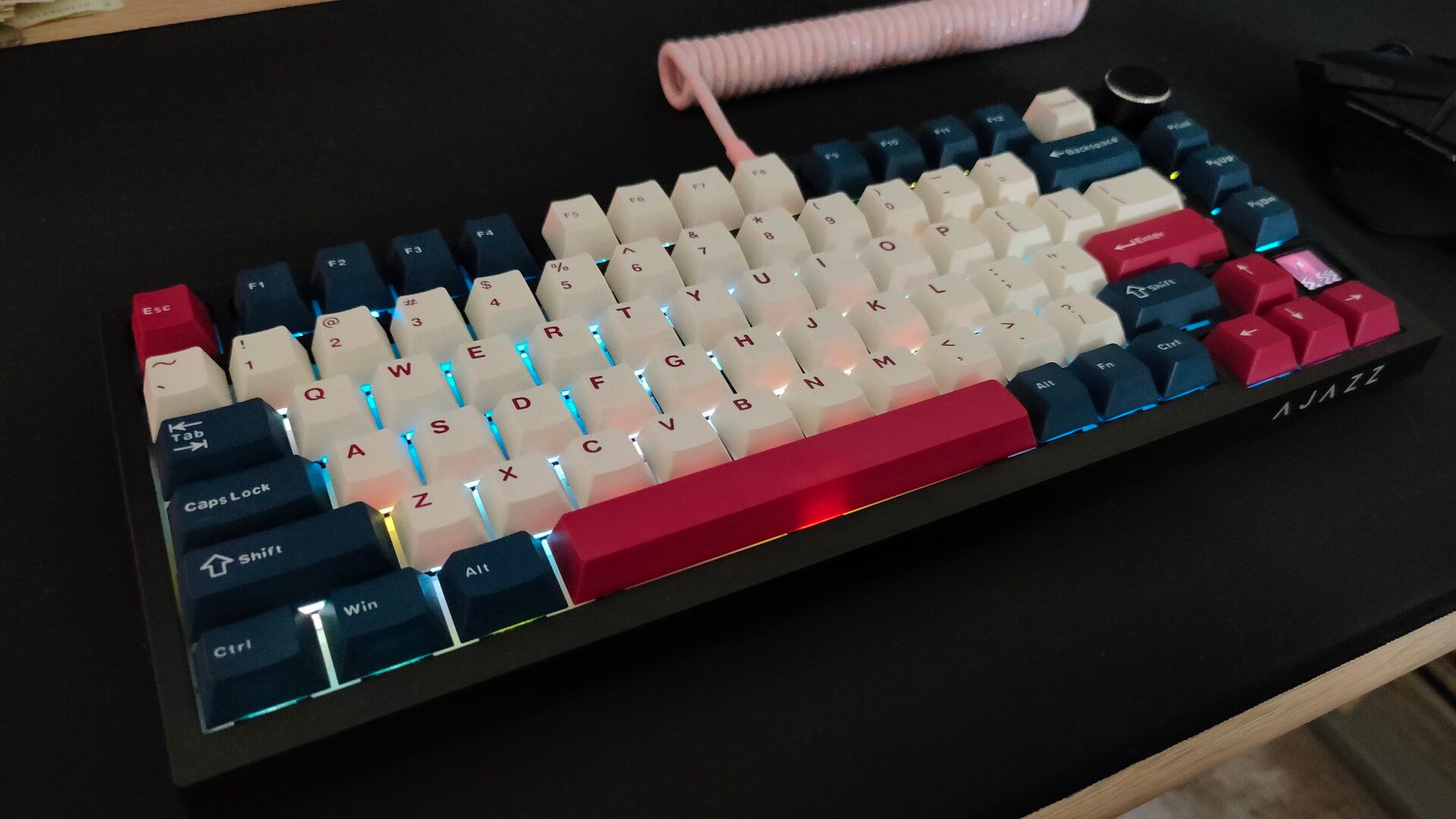 The Ajazz AK820 Pro mechanical keyboard with a pink coiled cable and white, blue, and red keycaps sitting on a desk with a black SteelSeries mousepad.