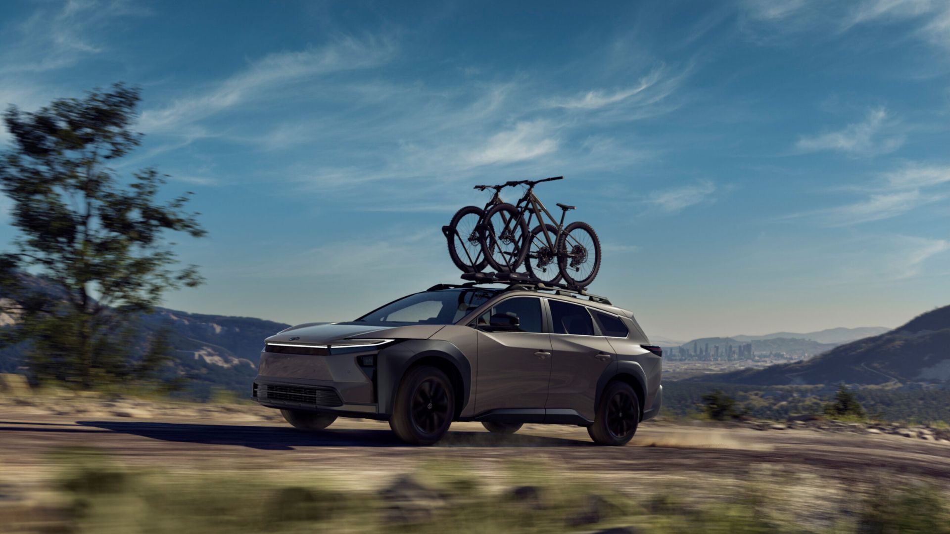 Dynamic front 3/4 shot of a 2026 Toyota bZ Woodland with mountain bikes on its roof driving on a dirt road with mountains in the background.